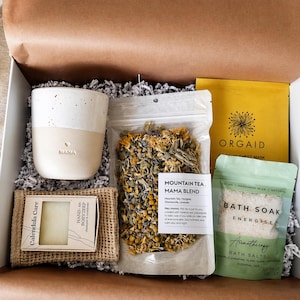 Op de afbeelding: Een cadeaupakket met zelfzorgartikelen. Inclusief een gespikkelde keramische mok met "MAMA", een zeep, een zakje thee, een sheetmasker en badzout. De doos is gevuld met versnipperd papier en bevat een handgeschreven briefje.