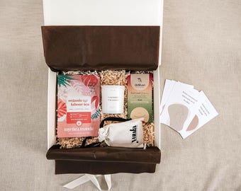 Può includere: Una scatola regalo bianca foderata con carta velina marrone scuro contenente una bustina di tè biologico Myrtle & Maude, una candela bianca etichettata A Moment for Mama, una tavoletta di cioccolato Delikao, un sacchetto beige con coulisse Yumla e carte.