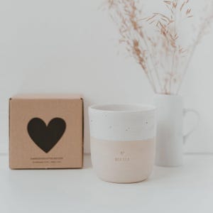 Pu&ograve; includere: Una tazza in ceramica bicolore con una parte superiore bianca maculata e una parte inferiore beige liscia. La parola MAMA e un piccolo cuore sono impressi sul davanti. Accanto si trova una scatola di cartone quadrata con un grande cuore nero.