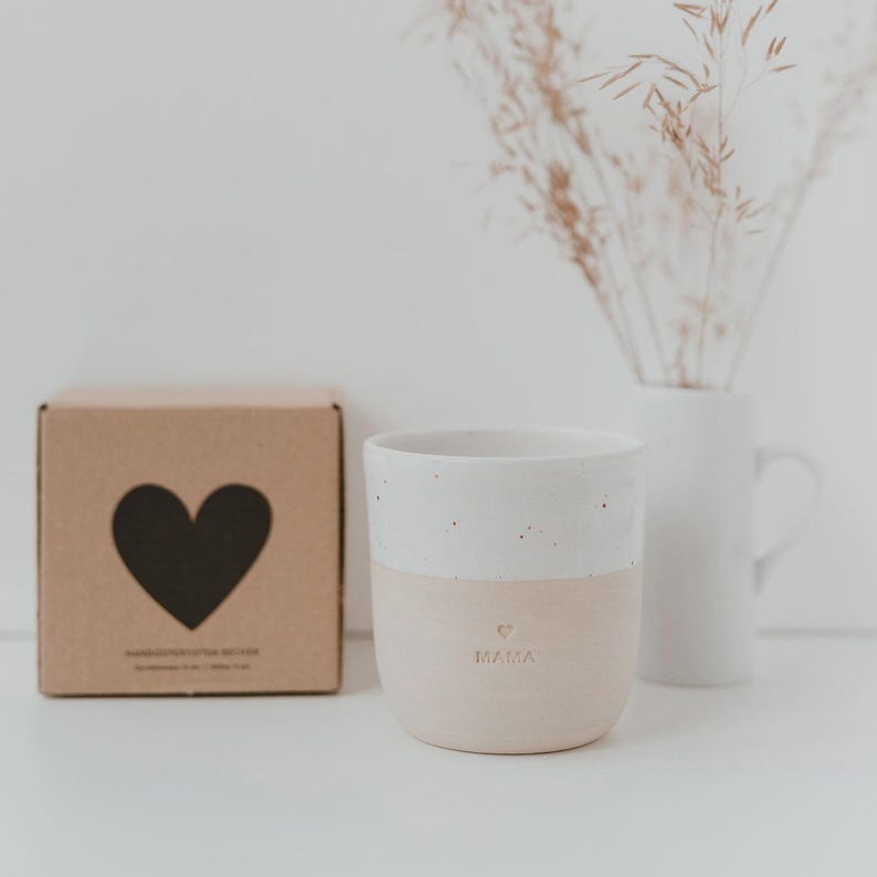 Pu&ograve; includere: Una tazza in ceramica bicolore con una parte superiore bianca maculata e una parte inferiore beige liscia. La parola MAMA e un piccolo cuore sono impressi sul davanti. Accanto si trova una scatola di cartone quadrata con un grande cuore nero.