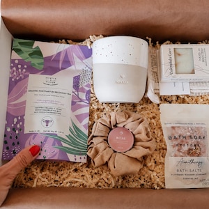 Op de afbeelding: Een geschenkdoos met een paars zakje Myrtle & Maude biologische thee voor postpartum herstel, een witte gespikkelde keramische mok met het woord MAMA, een stuk Calendula Care zeep, een beige stoffen scrunchie met een roze blikje en badzout.