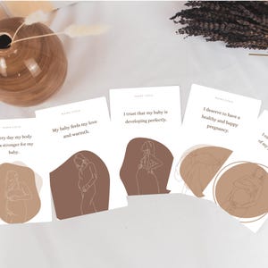 Op de afbeelding: Een verzameling witte kaarten met tekst en illustraties, met zinnen als "Mijn baby voelt mijn liefde en warmte" en "Ik verdien een gezonde en gelukkige zwangerschap". De kaarten hebben een minimalistisch ontwerp met bruine accenten en lijntekeningen van zwangere vrouwen.