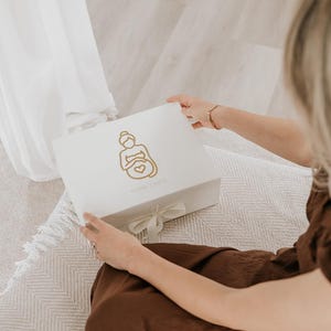 Pu&ograve; includere: Una scatola regalo rettangolare bianca con un'illustrazione a linee dorate di una persona incinta che tiene la pancia con un simbolo a forma di cuore. La scatola &egrave; chiusa con un fiocco di nastro bianco e presenta una piccola scritta dorata.
