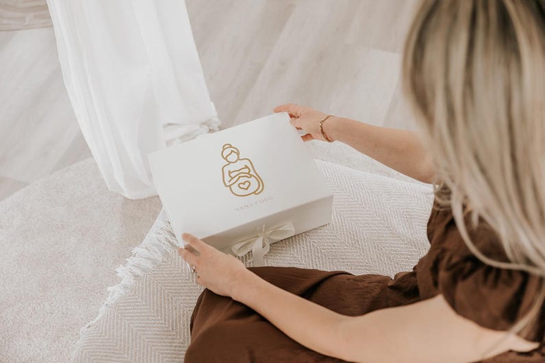 Pu&ograve; includere: Una scatola regalo rettangolare bianca con un'illustrazione a linee dorate di una persona incinta che tiene la pancia con un simbolo a forma di cuore. La scatola &egrave; chiusa con un fiocco di nastro bianco e presenta una piccola scritta dorata.