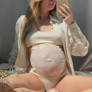Pu&ograve; includere: Una maschera in tessuto rotonda, bianca e testurizzata applicata su un ventre in gravidanza. La maschera presenta un bordo arricciato e un design circolare che copre l'addome. La persona indossa un top corto a coste beige, slip abbinati e una camicia aperta.
