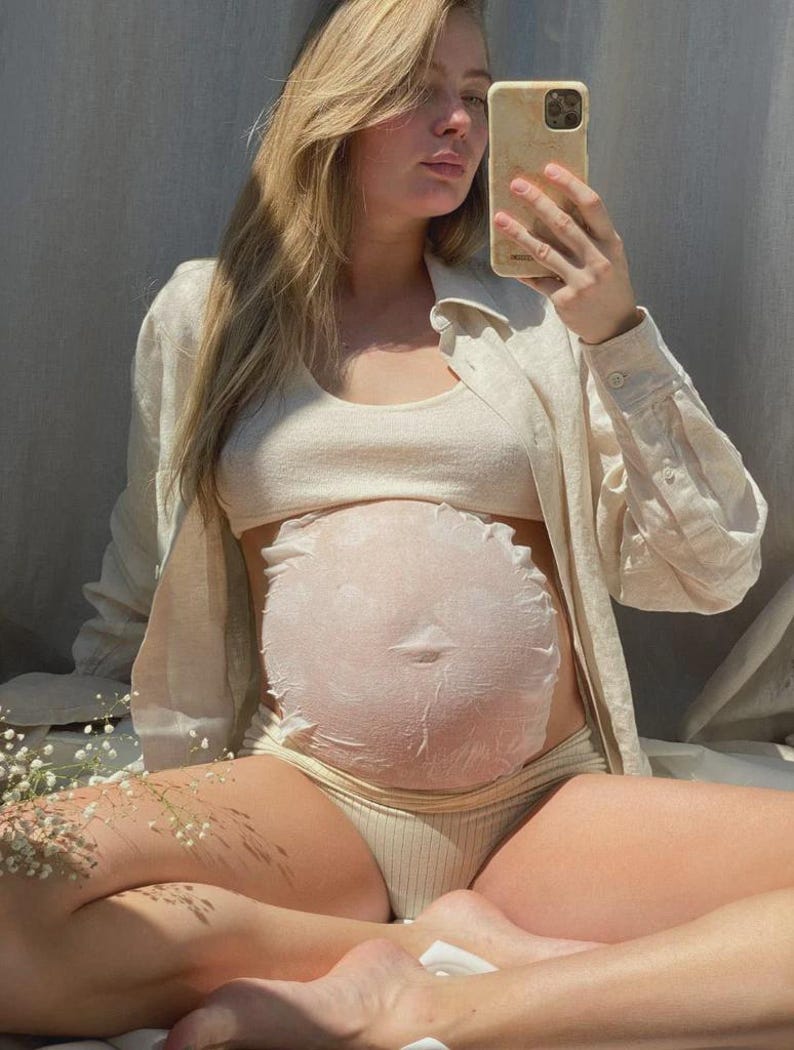 Pu&ograve; includere: Una maschera in tessuto rotonda, bianca e testurizzata applicata su un ventre in gravidanza. La maschera presenta un bordo arricciato e un design circolare che copre l'addome. La persona indossa un top corto a coste beige, slip abbinati e una camicia aperta.