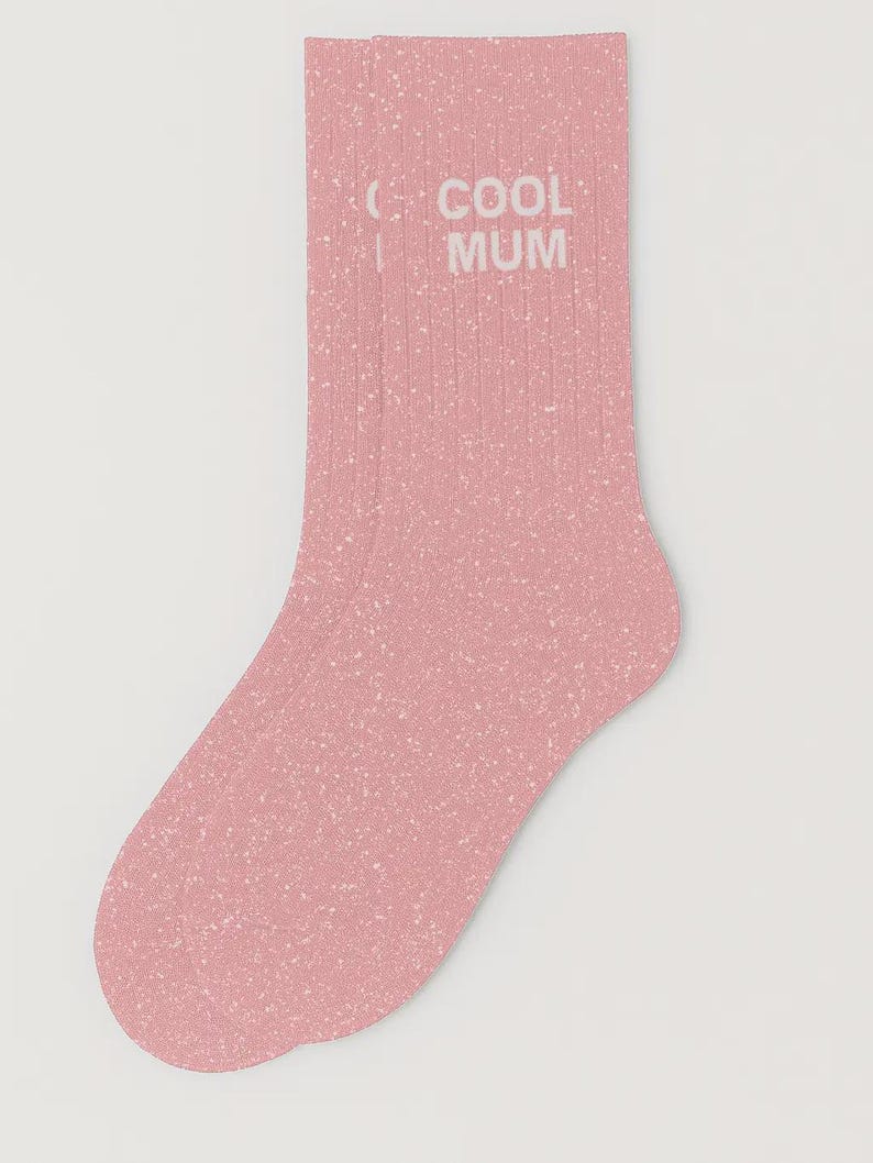 Pu&ograve; includere: Un paio di calzini a coste color rosa polvere con un motivo a macchie bianche su tutto il tessuto. Il testo COOL MUM &egrave; stampato in lettere maiuscole bianche sulla parte superiore dei calzini, posizionato verticalmente.