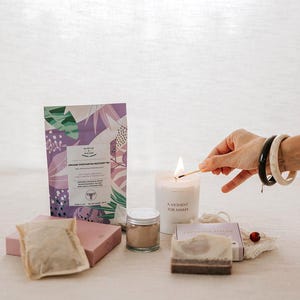 Pu&ograve; includere: Un set regalo benessere che include una busta di tisana biologica per il recupero post-partum con motivi botanici viola e verdi, una candela bianca con l'etichetta A MOMENT FOR MAMA, un vasetto di polvere, una saponetta e una bustina di t&egrave;.
