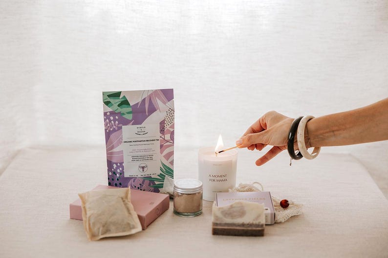 Pu&ograve; includere: Un set regalo benessere che include una busta di tisana biologica per il recupero post-partum con motivi botanici viola e verdi, una candela bianca con l'etichetta A MOMENT FOR MAMA, un vasetto di polvere, una saponetta e una bustina di t&egrave;.