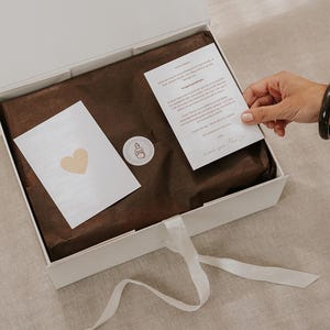 Pu&ograve; includere: Una scatola regalo rettangolare bianca con chiusura a nastro bianco, contenente carta velina marrone scuro, un piccolo adesivo circolare con un disegno a linee, un biglietto bianco con un cuore dorato e un biglietto con testo stampato.