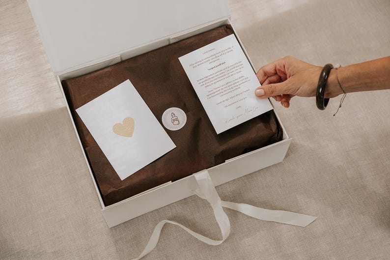 Pu&ograve; includere: Una scatola regalo rettangolare bianca con chiusura a nastro bianco, contenente carta velina marrone scuro, un piccolo adesivo circolare con un disegno a linee, un biglietto bianco con un cuore dorato e un biglietto con testo stampato.