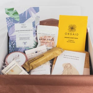 Op de afbeelding: Een geschenkdoos gevuld met zelfzorgartikelen. Inclusief een zakje thee, een geel gezichtsmasker, badzout, een zeep, een magnesiumsupplement en een kaart met tekst. De doos is wit met een bruin interieur.