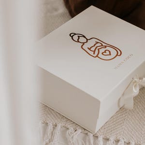 Pu&ograve; includere: Una scatola regalo rettangolare bianca con coperchio incernierato e chiusura a nastro bianco. La parte superiore della scatola presenta un'illustrazione a linee marroni di una persona con un cuore sull'addome e la scritta MAMA COCO in marrone.