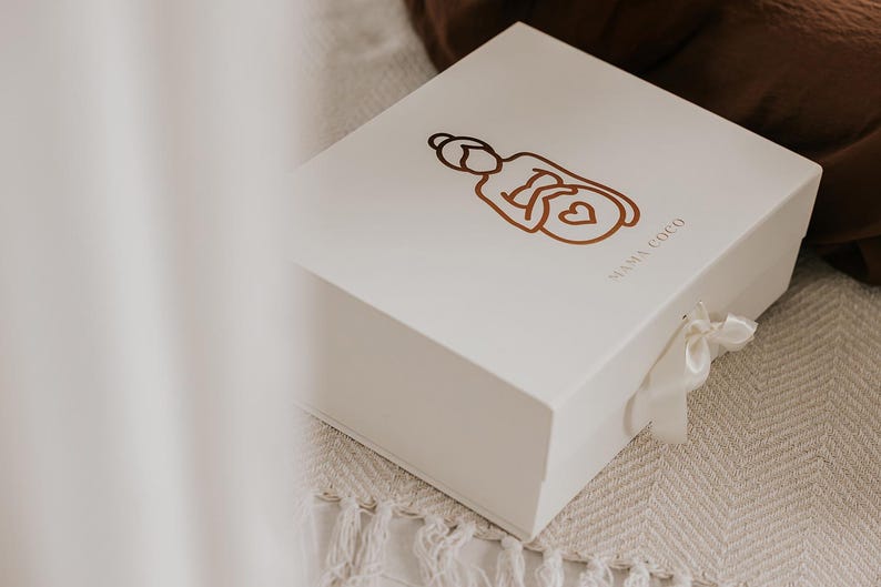 Pu&ograve; includere: Una scatola regalo rettangolare bianca con coperchio incernierato e chiusura a nastro bianco. La parte superiore della scatola presenta un'illustrazione a linee marroni di una persona con un cuore sull'addome e la scritta MAMA COCO in marrone.