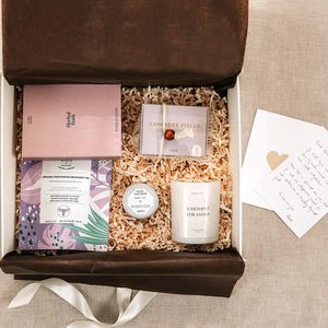 Pu&ograve; includere: Una scatola regalo riempita con carta sminuzzata contenente una scatola rosa Herbal Bath, un pacchetto Lavender Fields con una perlina rossa, un piccolo barattolo rotondo, una candela bianca con etichetta A Moment for Mama, una bustina di t&egrave; viola e un biglietto.