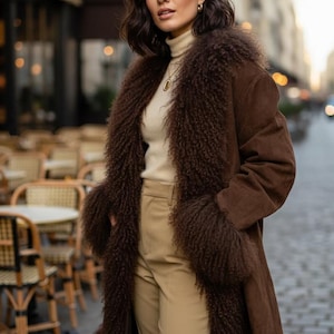 May include: A long, brown coat with a faux fur collar and cuffs. The coat is worn over a cream-coloured turtleneck and tan trousers. The woman is standing on a cobbled street.
