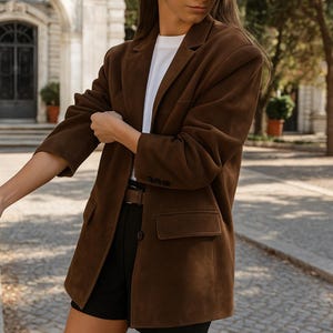 Puede incluir: Un blazer de ante marrón con un diseño clásico. El blazer tiene solapas con muescas, dos bolsillos delanteros y tres botones en los puños. La modelo lleva una camiseta blanca y pantalones cortos negros. El blazer es de color marrón medio.
