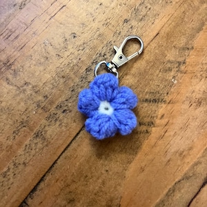 Puede incluir: Un llavero de flor azul tejido a ganchillo con un centro blanco. La flor está unida a un mosquetón y un llavero de metal plateado. El fondo es una superficie de madera.
