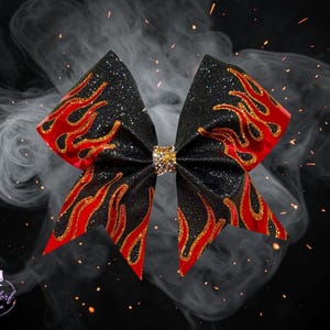 May include: A black glitter hair bow featuring a red and gold flame pattern across the fabric. The center of the bow is cinched with a band of small, reflective gold-toned crystals. The design is symmetrical with pointed ends on each side.