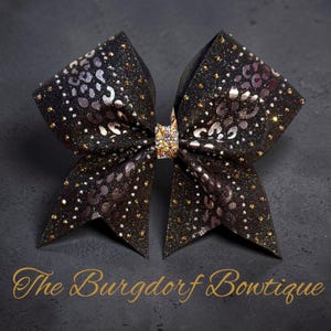 May include: A black and gold glitter bow with a leopard print design. The bow has a rhinestone center and is decorated with gold glitter. The text "The Burgdorf Bowtique" is at the bottom.