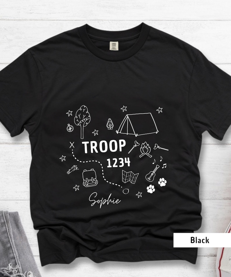 K&ouml;nnte beinhalten: Schwarzes T-Shirt mit wei&szlig;em Camping-Motiv. Das Design umfasst ein Zelt, einen Baum, ein Lagerfeuer, eine Karte, eine Gitarre, einen Rucksack und Pfotenabdr&uuml;cke. Der Text lautet "TROOP 1234 Sophie".