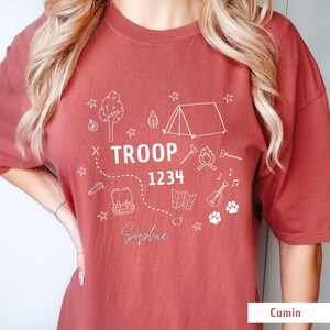 May include: Cumin-colored t-shirt with a white graphic design featuring camping-themed illustrations. The design includes a tent, tree, campfire, map, backpack, guitar, and paw prints. The text "TROOP 1234" and the name "Sophie" are also included.