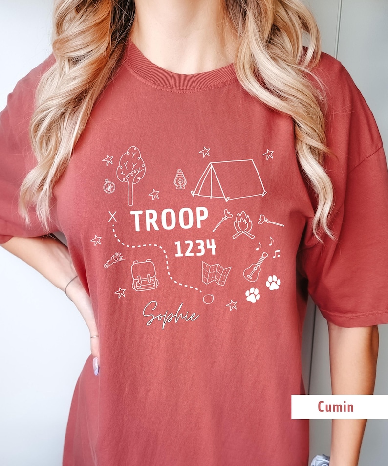 May include: Cumin-colored t-shirt with a white graphic design featuring camping-themed illustrations. The design includes a tent, tree, campfire, map, backpack, guitar, and paw prints. The text "TROOP 1234" and the name "Sophie" are also included.