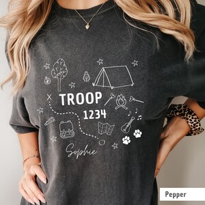May include: Dark gray t-shirt with white camping-themed graphics, including a tent, campfire, and map. The text "TROOP 1234 Sophie" is displayed in the center. The shirt is a comfortable, casual style.