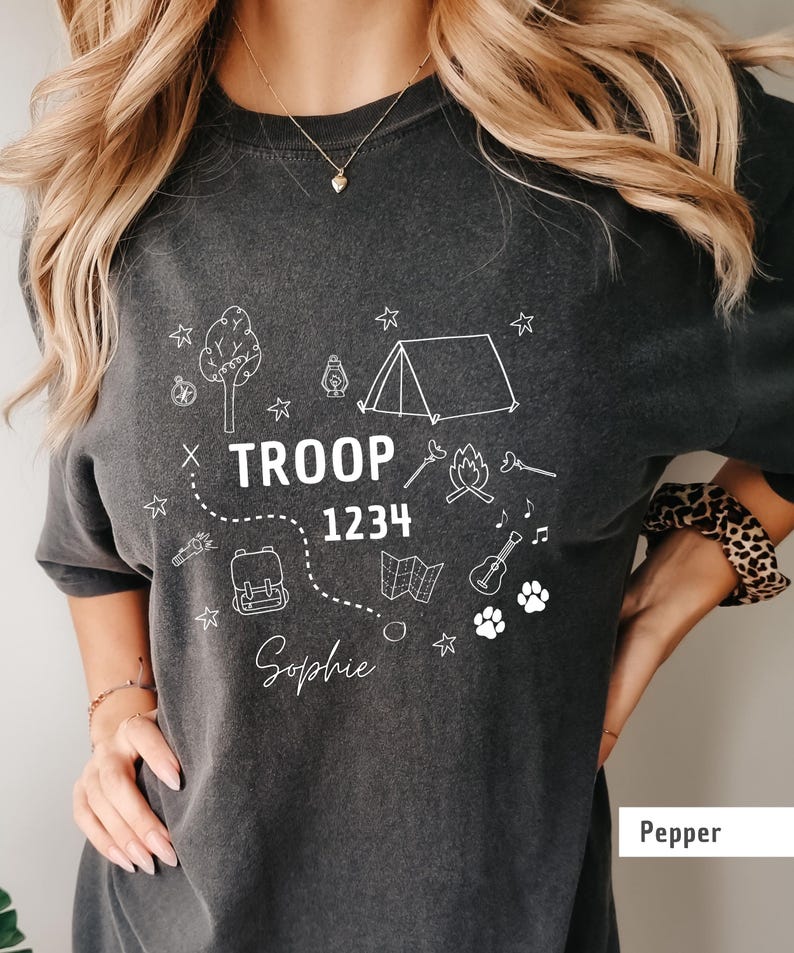 May include: Dark gray t-shirt with white camping-themed graphics, including a tent, campfire, and map. The text "TROOP 1234 Sophie" is displayed in the center. The shirt is a comfortable, casual style.