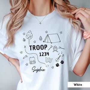 May include: White t-shirt with a black graphic design featuring camping-themed illustrations and the text "TROOP 1234 Sophie." The design includes a tent, campfire, backpack, map, guitar, paw prints, and stars.