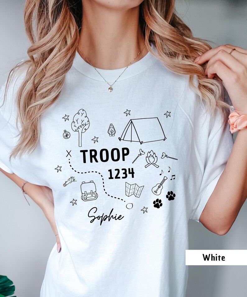 May include: White t-shirt with a black graphic design featuring camping-themed illustrations and the text "TROOP 1234 Sophie." The design includes a tent, campfire, backpack, map, guitar, paw prints, and stars.