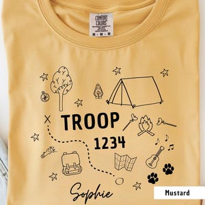 K&ouml;nnte beinhalten: Senffarbenes T-Shirt mit Rundhalsausschnitt. Das Shirt hat eine schwarze Grafik mit Camping-Elementen, darunter ein Zelt, ein Lagerfeuer und eine Karte. Der Text enth&auml;lt "TROOP 1234" und "Sophie."