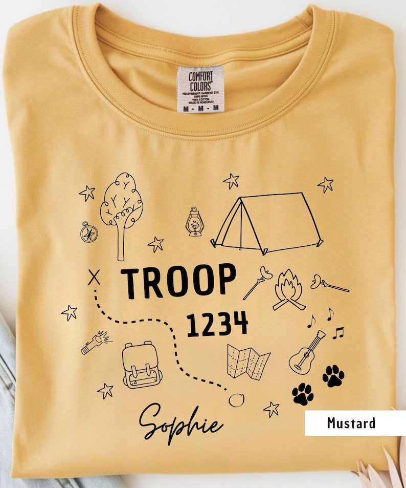 K&ouml;nnte beinhalten: Senffarbenes T-Shirt mit Rundhalsausschnitt. Das Shirt hat eine schwarze Grafik mit Camping-Elementen, darunter ein Zelt, ein Lagerfeuer und eine Karte. Der Text enth&auml;lt "TROOP 1234" und "Sophie."
