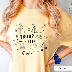 May include: A light yellow t-shirt with a black graphic design featuring camping-themed illustrations, including a tent, campfire, and map. The text "TROOP 1234 Sophie" is also printed on the shirt.