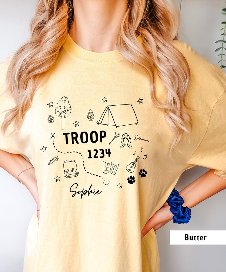 May include: A light yellow t-shirt with a black graphic design featuring camping-themed illustrations, including a tent, campfire, and map. The text "TROOP 1234 Sophie" is also printed on the shirt.