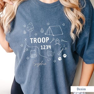 May include: Denim blue t-shirt with white graphic illustrations of camping elements, including a tent, tree, campfire, and map. The text "TROOP 1234" and the name "Sophie" are also printed on the shirt.