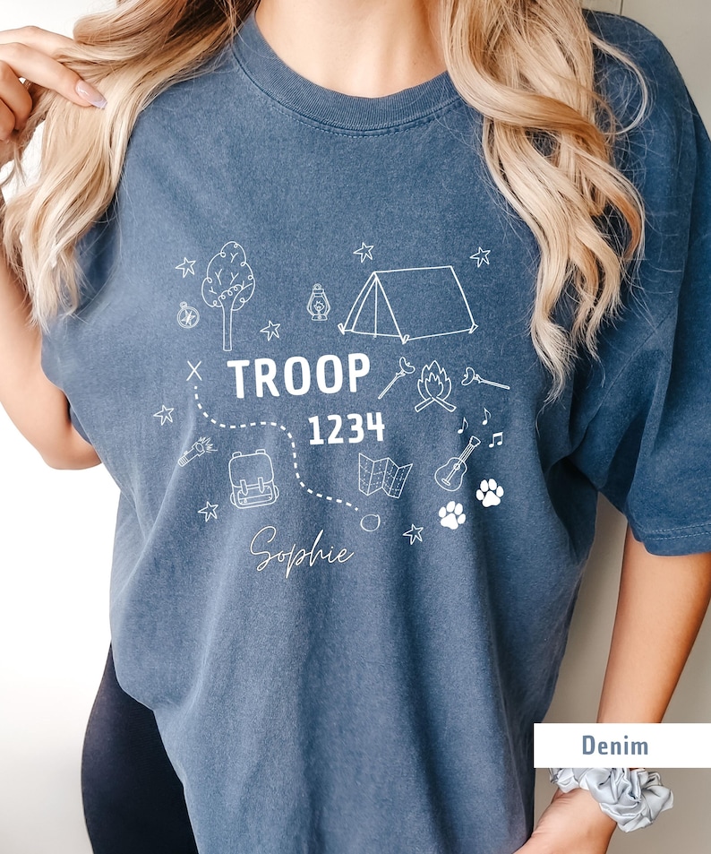 May include: Denim blue t-shirt with white graphic illustrations of camping elements, including a tent, tree, campfire, and map. The text "TROOP 1234" and the name "Sophie" are also printed on the shirt.
