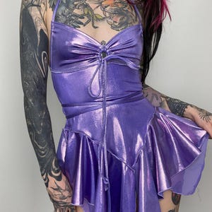 May include: A shimmering purple halter dress with a cinched waist and a layered, ruffled skirt. The dress features a tie detail at the bust and a flowing, asymmetrical hemline. The fabric has a metallic sheen, catching the light.
