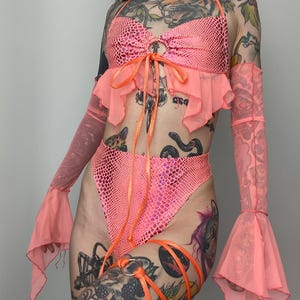 May include: A neon pink two-piece outfit with a halter top, high-waisted bottoms, and matching arm sleeves. The top and bottoms have a textured pattern, and the outfit is accented with orange ties and ruffles. A unique and eye-catching ensemble.