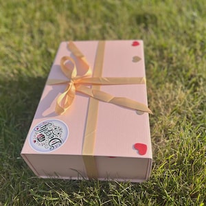 Puede incluir: Una caja de regalo rosa pálido con una cinta dorada atada en un lazo. La caja tiene una pegatina con la palabra "Deeply" y pequeñas pegatinas en forma de corazón. La caja está sobre hierba verde.