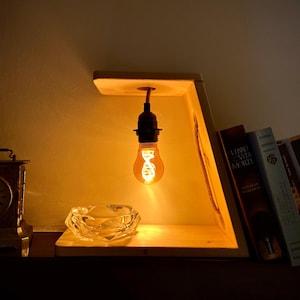 Puede incluir: Una lámpara de madera con una bombilla colgante ilumina un cenicero de cristal claro y facetado. Un reloj vintage y varios libros también son visibles. La luz cálida crea un ambiente acogedor.