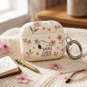 May include: Off-white AirPods case decorated with pink floral and Snoopy illustrations. The case has a silver-colored ring for attaching to a keychain. A small notebook, pen, and glasses are also in the image.