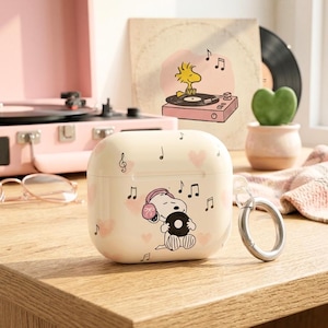 May include: Off-white AirPods case featuring a Snoopy graphic with a record and musical notes. The case has a silver metal ring and is decorated with pink hearts. A pink record player and other decorative items are in the background.