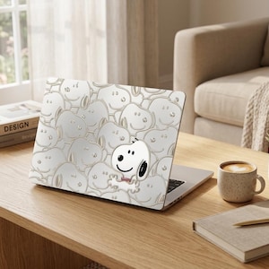 Op de afbeelding: Een laptop met een Snoopy-ontwerp op de klep staat op een houten tafel. Het ontwerp toont een herhalend patroon van Snoopy's gezicht in lichtgrijs, met een grotere Snoopy in het midden. Een kopje koffie, een notitieboek en een vetplant staan ook op de tafel.
