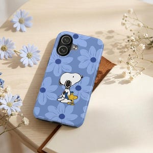 Puede incluir: Funda de teléfono azul con un estampado floral y una imagen de dibujos animados de Snoopy y Woodstock. La funda está diseñada para proteger el teléfono de arañazos e impactos. Acabado brillante.