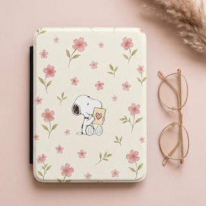 Op de afbeelding: Een crèmekleurige e-readerhoes met een herhalend patroon van kleine roze bloemen met groene stengels en bladeren. In het midden staat een getekende hond afgebeeld die zit en een klein beige kaartje met een roze hartje vasthoudt.