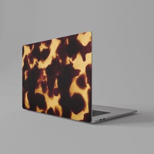 May include: Laptop with a tortoiseshell pattern on the lid. The design features a mix of dark brown and golden-yellow hues, creating a marbled effect. The laptop is silver and sits on a gray surface.