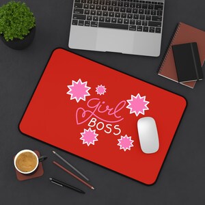 May include: A red rectangular mousepad with the words "Girl Boss" in a cursive font, accompanied by pink starburst designs. A white computer mouse is on the right side of the mousepad. A laptop and other office supplies are in the background.