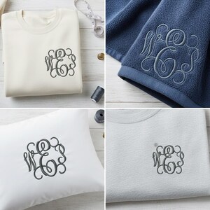 Op de afbeelding: Een verzameling gepersonaliseerde items, waaronder een cr&egrave;mekleurige sweatshirt, een marineblauwe handdoek, een wit kussen en een lichtgrijs t-shirt. Elk item heeft een donkergrijs geborduurd monogramontwerp. De items zijn gerangschikt op een licht houten oppervlak.