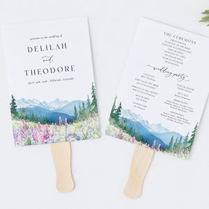 May include: Two white paper wedding fans with wooden handles. The fans feature a watercolor design of mountains, trees, and wildflowers. One fan displays the names Delilah and Theodore, the other lists the ceremony details.